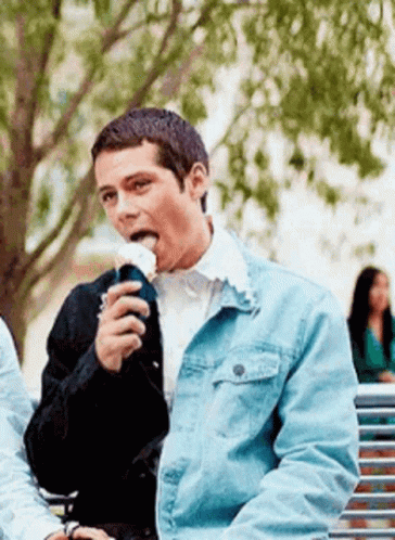 A Man In A Denim Jacket Is Sitting On A Park Bench Eating A Sandwich . GIF