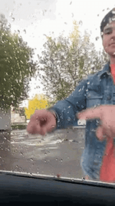 A Man In A Denim Jacket Is Standing In Front Of A Car Windshield With Rain Drops On It . GIF