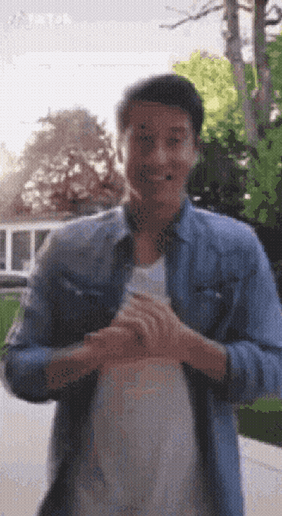 A Man In A Denim Shirt And White Shirt Is Standing In Front Of A Tree . GIF