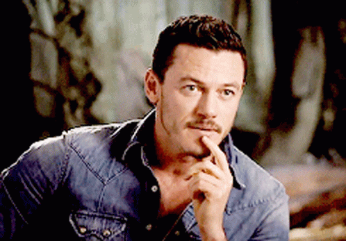 A Man In A Denim Shirt Holds His Hand To His Chin And Looks At The Camera GIF