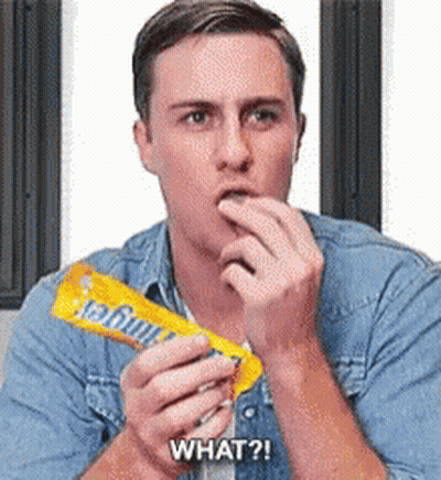 A Man In A Denim Shirt Is Eating A Candy Bar And Saying What ? GIF