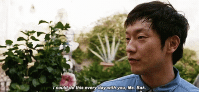 A Man In A Denim Shirt Is Talking To Another Man In Front Of A Garden . GIF