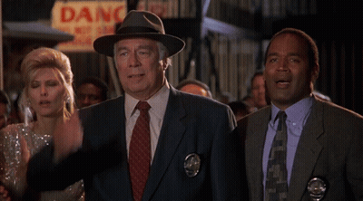 A Man In A Fedora Stands In Front Of A Sign That Says Danger GIF