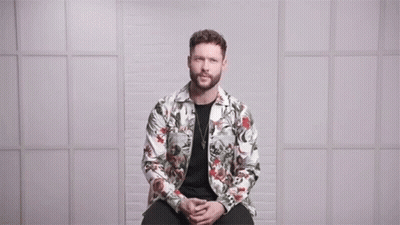 A Man In A Floral Jacket Is Sitting In Front Of A Wall With The Words Love Conquers All Above Him GIF