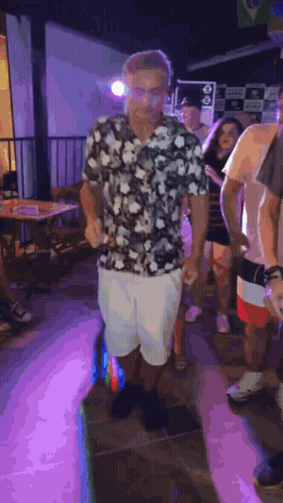 A Man In A Floral Shirt And White Shorts Is Dancing In A Crowd Of People At A Party . GIF