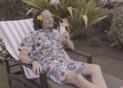 A Man In A Floral Shirt Is Sitting In A Chair Drinking A Cocktail GIF