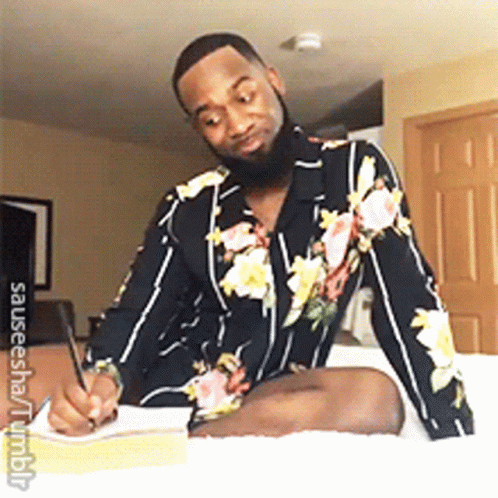 A Man In A Floral Shirt Is Sitting On A Bed Writing In A Notebook GIF