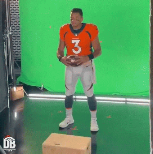 A Man In A Football Uniform Is Standing On A Box Holding A Football In Front Of A Green Screen . GIF