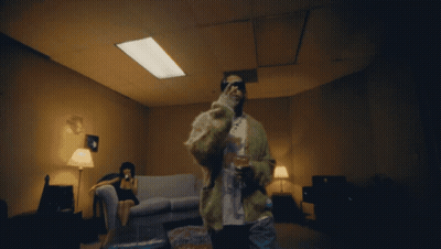 A Man In A Fur Coat Stands In A Dark Room With A Woman Sitting On A Couch GIF