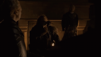 A Man In A Fur Coat Stands In A Dark Room With Two Other People GIF