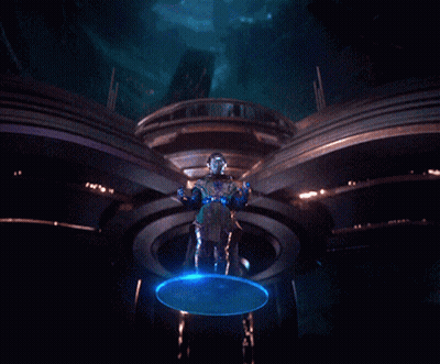 A Man In A Futuristic Outfit Is Standing In A Dark Room With A Blue Light Behind Him GIF