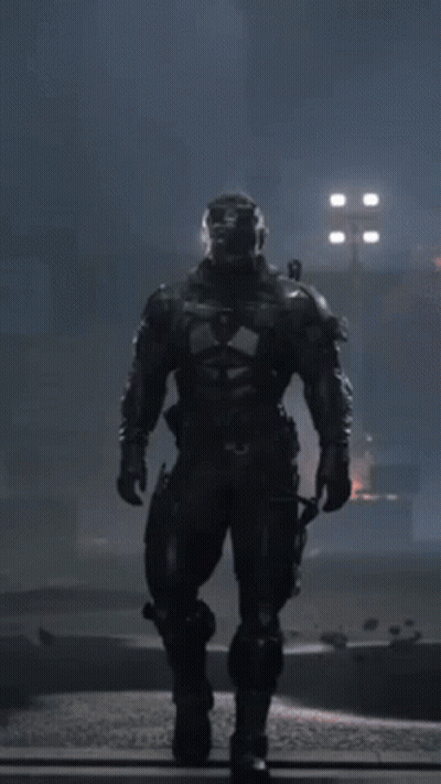 A Man In A Futuristic Suit With A Helmet On Stands In The Dark GIF