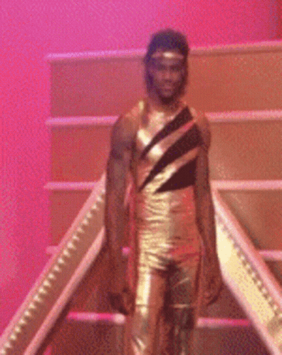 A Man In A Gold Leotard Is Standing On A Set Of Stairs GIF