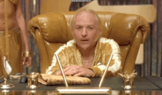 A Man In A Gold Robe Is Sitting At A Desk With A Pen . GIF