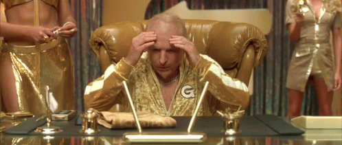 A Man In A Gold Robe With The Letter G On His Chest Is Sitting At A Desk . GIF