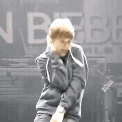 A Man In A Gray Jacket Is Dancing On A Stage In Front Of A Sign That Says Justin Bieber . GIF