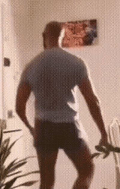 A Man In A Gray Shirt And Black Shorts Is Standing In A Room With A Picture On The Wall Behind Him . GIF