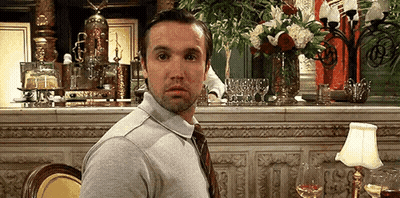 A Man In A Gray Shirt And Tie Is Sitting At A Table In A Restaurant With A Vase Of Flowers In The Background . GIF