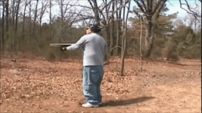 A Man In A Gray Shirt Is Holding A Shotgun In A Field GIF