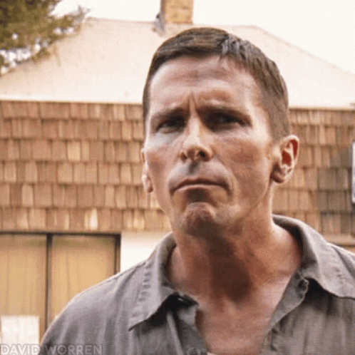 A Man In A Gray Shirt Is Standing In Front Of A Building With A Roof Made Of Wood Shingles . GIF