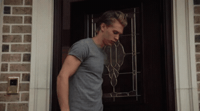 A Man In A Gray Shirt Is Standing In Front Of A Door GIF