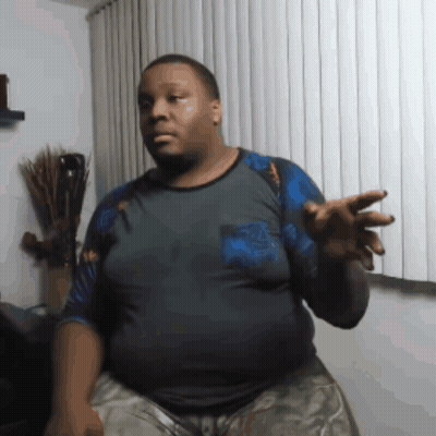 A Man In A Gray Shirt Is Standing In Front Of A Window With Blinds GIF
