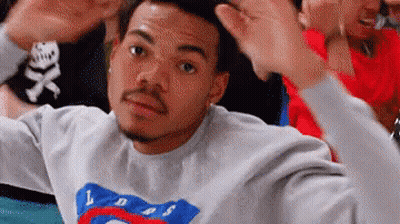 A Man In A Gray Sweatshirt Is Making A Funny Face While Sitting In A Crowd Of People . GIF