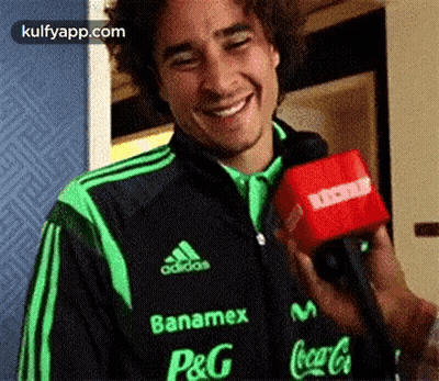 A Man In A Green And Black Adidas Jacket Is Holding A Red Microphone And Smiling . GIF