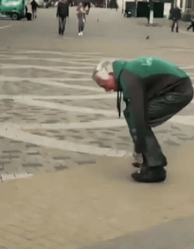 A Man In A Green And Black Jacket Is Tying His Shoes On A Sidewalk GIF