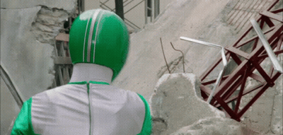 A Man In A Green And White Costume Is Standing In Front Of A Destroyed Building GIF