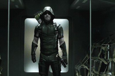A Man In A Green Arrow Costume Is Standing In A Dark Room Holding A Bow And Arrow . GIF