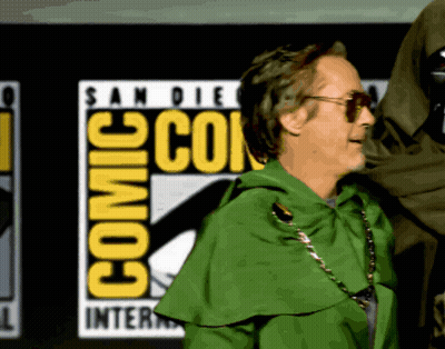 A Man In A Green Cape Is Standing In Front Of A Comic Con Logo GIF