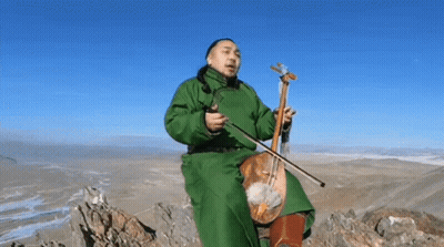 A Man In A Green Coat Is Sitting On Top Of A Mountain Playing A Violin . GIF
