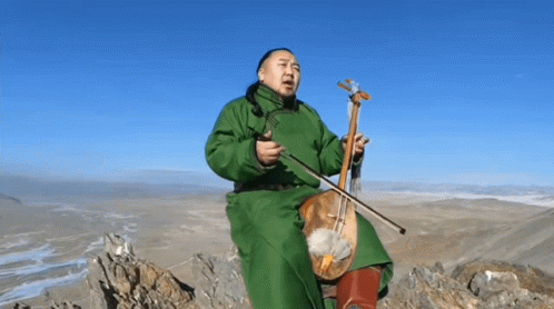 A Man In A Green Coat Sits On A Rock Holding A Violin GIF