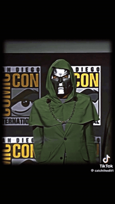 A Man In A Green Hooded Jacket Stands In Front Of A Comic Con Poster GIF