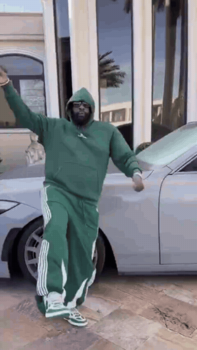 A Man In A Green Hoodie And Green Pants Is Standing In Front Of A Silver Car . GIF