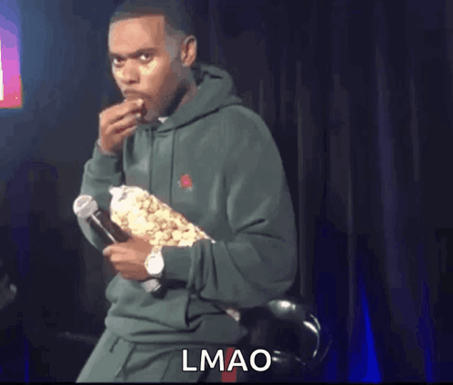 A Man In A Green Hoodie Is Holding A Microphone And Eating Popcorn . GIF