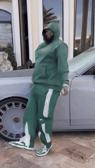 A Man In A Green Hoodie Is Standing In Front Of A Silver Car GIF