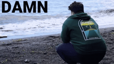 A Man In A Green Hoodie Squatting On A Beach With The Word Damn Written Above Him GIF