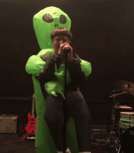 A Man In A Green Inflatable Alien Costume Singing Into A Microphone GIF
