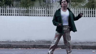 A Man In A Green Jacket And Camo Pants Is Dancing On A Street GIF