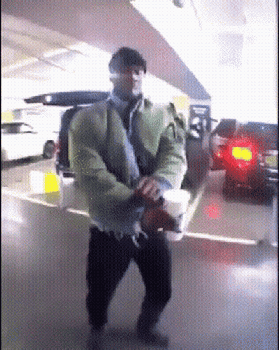 A Man In A Green Jacket Is Holding A Cup Of Coffee In A Parking Garage GIF