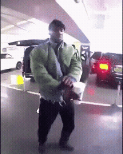 A Man In A Green Jacket Is Holding A Cup Of Coffee In A Parking Garage GIF