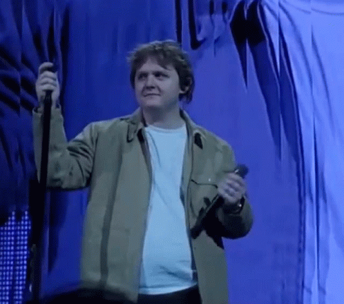 A Man In A Green Jacket Is Holding A Microphone On A Stage In Front Of A Blue Curtain . GIF