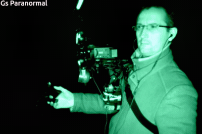 A Man In A Green Jacket Is Standing In A Dark Room With Gs Paranormal Written On The Bottom Right GIF