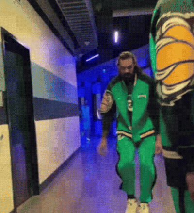 A Man In A Green Jacket Is Walking Down A Hallway With Another Man . GIF