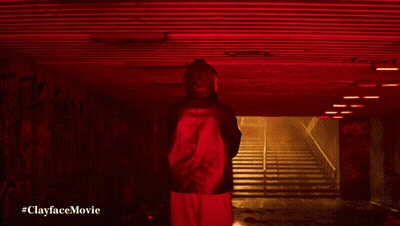 A Man In A Green Jacket Stands In A Dark Tunnel With The Words #Clayfacemovie Below Him GIF