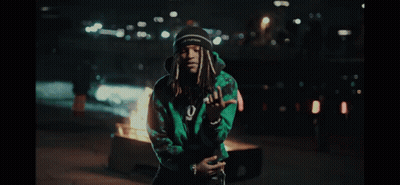A Man In A Green Jacket Stands In Front Of A Burning Trash Can GIF