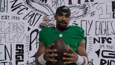 A Man In A Green Jersey Holds A Football In Front Of A Wall That Says Eagles Fly On The Road To Victory GIF