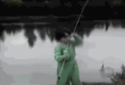 A Man In A Green Jumpsuit Is Holding A Fishing Rod In Front Of A Lake . GIF